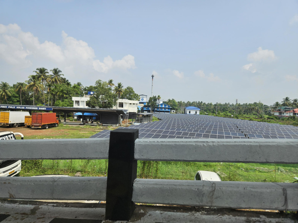 Großflächige Aufdach-Photovoltaikanlage auf einem Industriegebäude in Kerala. Im Hintergrund ist ein Schild mit der Aufschrift „First Fully Solar Powered Dairy" zu lesen; Palmen und tropische Vegetation umgeben das Gelände.