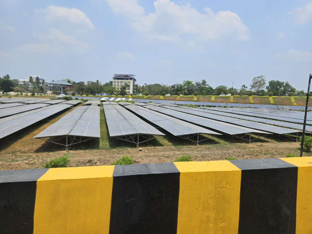 Weitläufige Freiflächen-Photovoltaikanlage in Kerala, Indien. Zahlreiche aufgeständerte Solarmodulreihen erstrecken sich unter blauem Himmel, im Hintergrund Palmen und Gebäude.