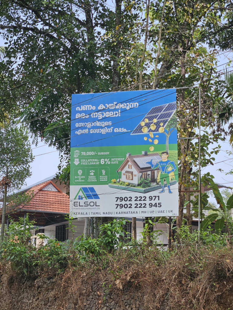 Werbeplakat des Solaranbieters Elsol Power Solutions im ländlichen Kerala, Indien. Das Schild auf Malayalam und Englisch bewirbt staatliche Subventionen (78.000 Rupien) und zinsgünstige Kredite für Haus-Solaranlagen; im Hintergrund ein traditionelles Wohnhaus umgeben von tropischer Vegetation.