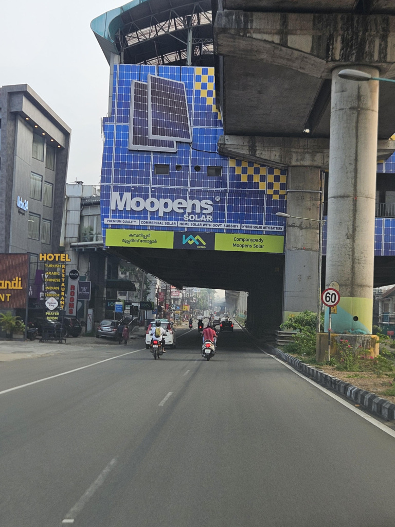 Werbefassade eines Solarhändlers (Moopens Solar) an einem Metrobahnhof in Kochi, Kerala. Die Fassade ist mit aufgedruckten und realen Solarmodulen gestaltet; darunter fahren Motorräder und Autos durch die belebte Stadtstraße.