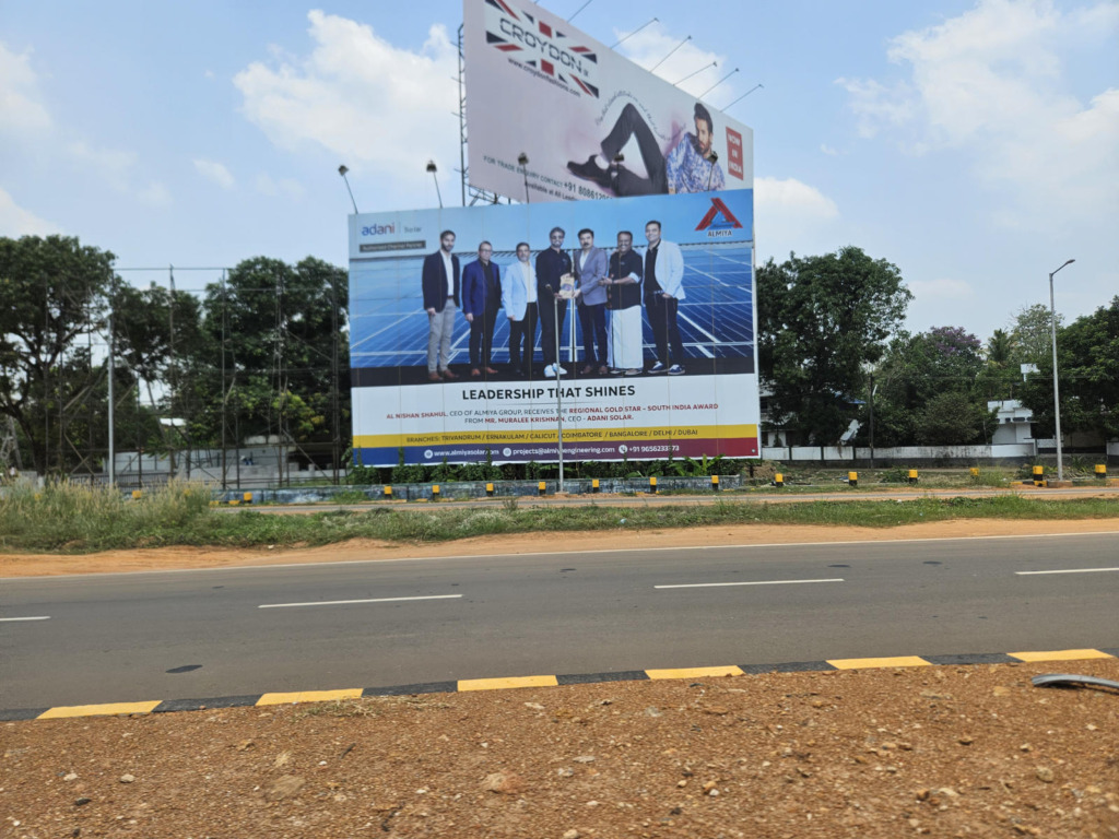 Großes Werbeplakat eines Solar-Unternehmens (Almiya/Adani Solar) an einer Straße in Kerala, Indien. Das Plakat zeigt eine Preisverleihung vor einer Solaranlage mit dem Slogan „Leadership That Shines", im Hintergrund tropische Vegetation.