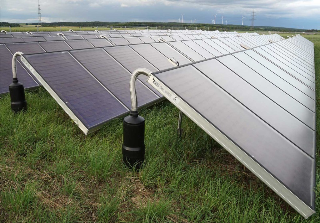 Großflächige Solarkollektoren einer Freiflächenanlage auf einer Wiese, mit sichtbaren Anschlussrohren und Absperrventilen. Im Hintergrund Windräder und Hochspannungsmasten unter bewölktem Himmel.