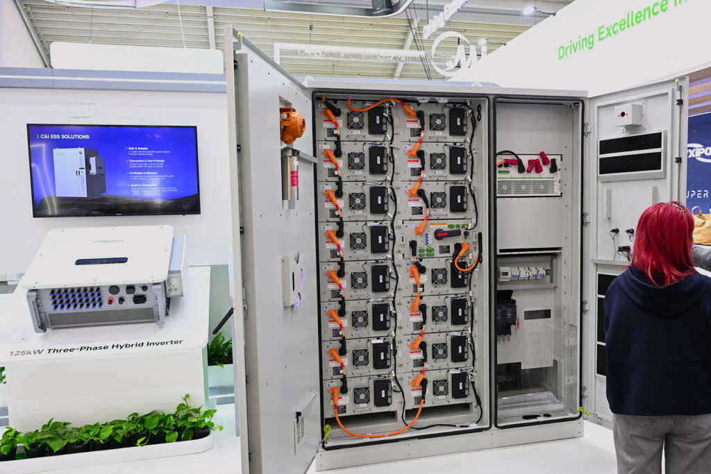 Geöffneter Schaltschrank eines gewerblichen Batteriespeichers mit Batteriemodule, orangefarbenen Hochvolt-Kabeln und Lüftern auf einer Energiemesse