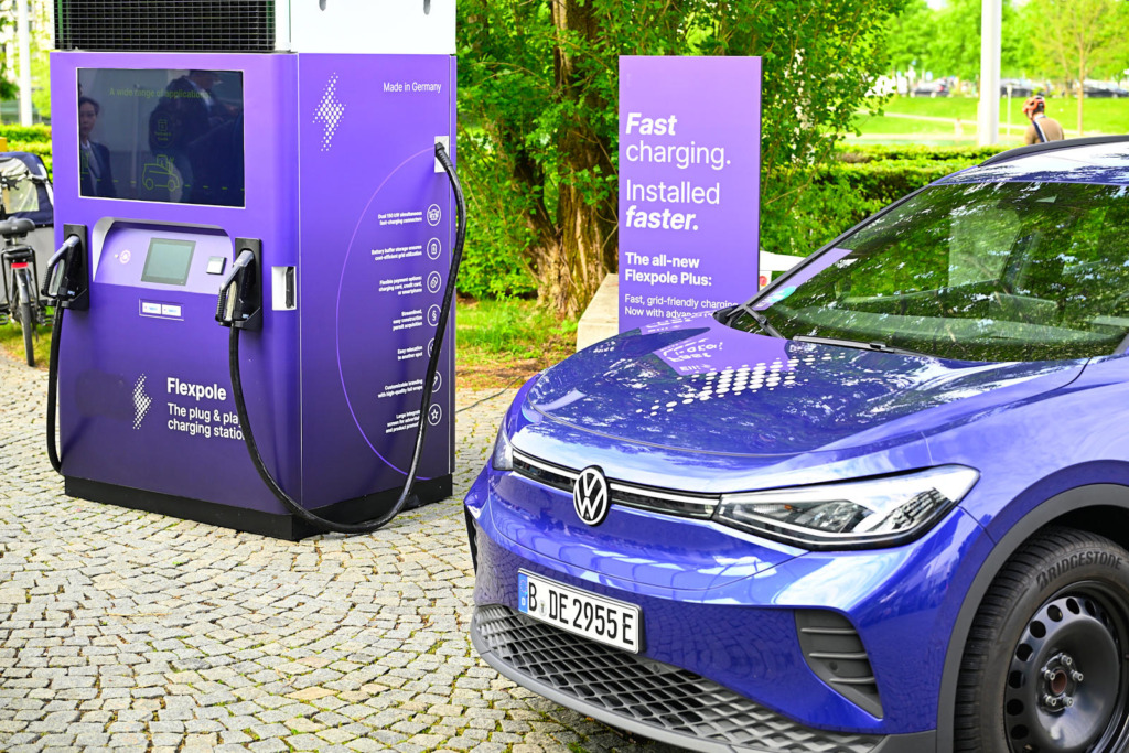 Lila Schnellladestation „Flexpole Plus" mit integriertem Batteriespeicher neben einem blauen VW ID.4 auf einem Pflasterplatz im Freien