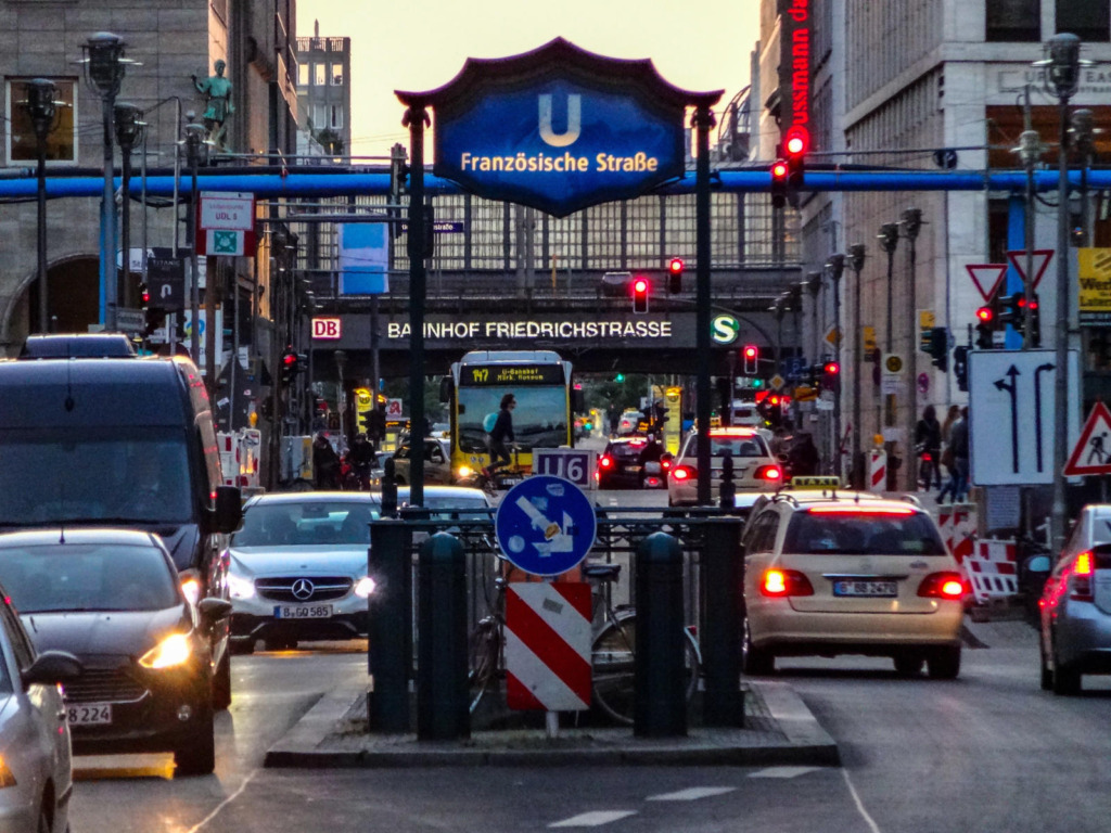 Belebte Stadtstraße in Berlin in der Abenddämmerung mit dichtem Fahrzeugverkehr. Im Zentrum des Bildes ist der U-Bahn-Eingang der Station „Französische Straße" (Linie U6) mit charakteristischem blauem Schild zu sehen. Im Hintergrund das Bahnhofsschild „Bahnhof Friedrichstraße" mit DB- und S-Bahn-Logo. Auf der Straße Baustellenabsperrungen und Verkehrsschilder.