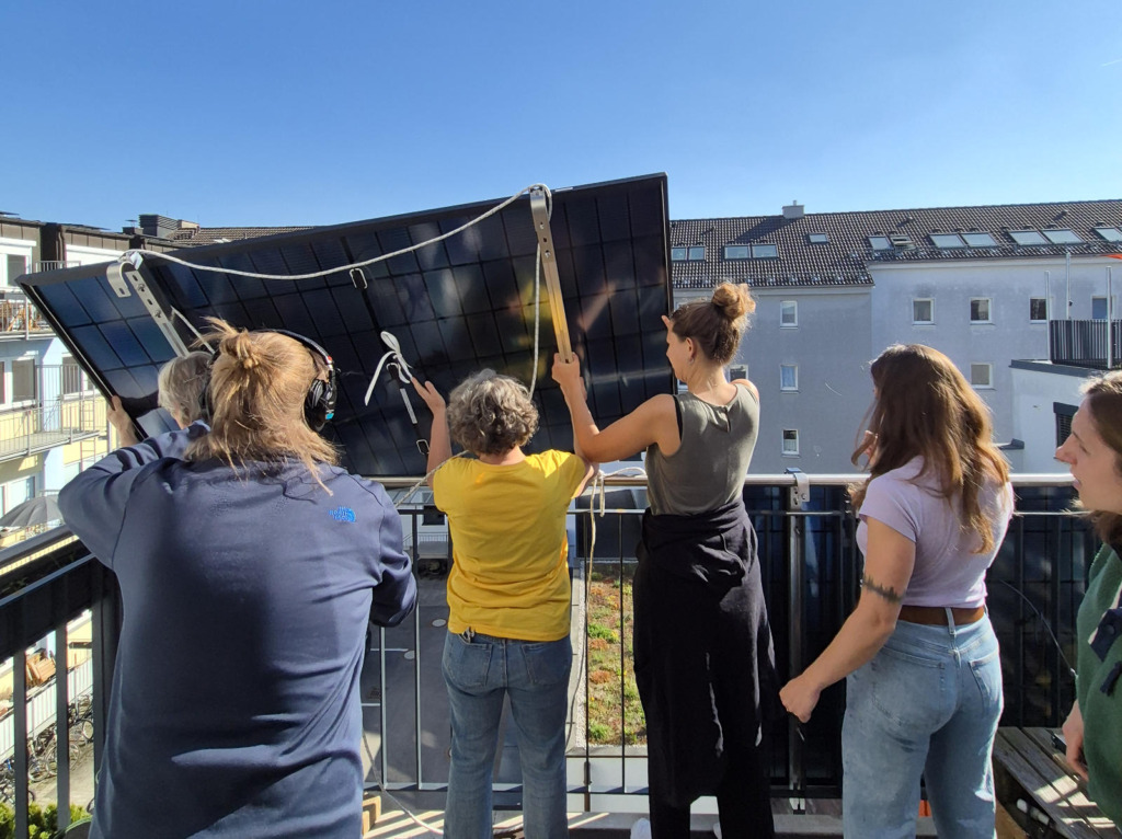 Mehrere Frauen installieren gemeinsam ein Balkonkraftwerk auf einem Balkon, indem sie das Solarmodul an der Balkonbrüstung befestigen.
