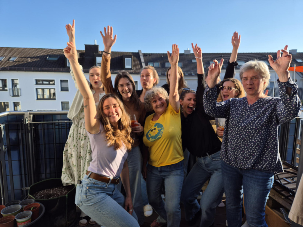 Gruppe von Frauen jubelt mit erhobenen Armen auf einem Balkon mit Solarmodul, im Hintergrund die Dächer der Nachbarhäuser bei Sonnenschein.