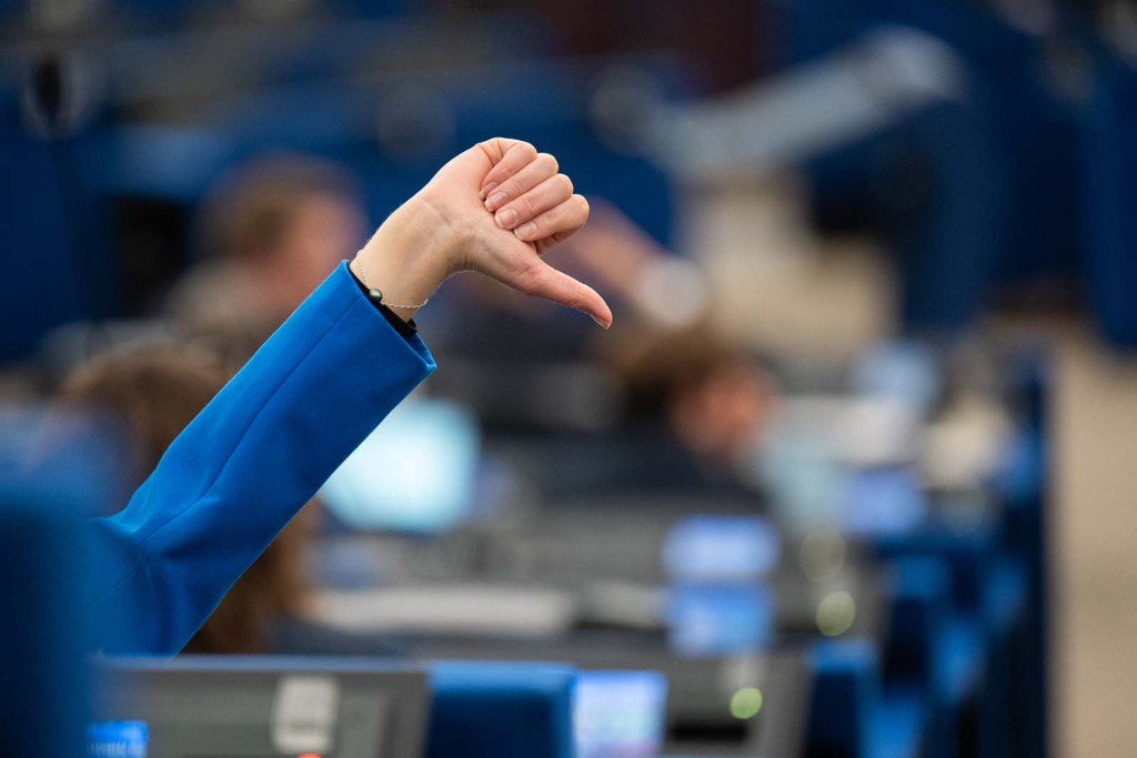 Erhobener Arm im EU-Parlament mit nach unten gerichtetem Daumen als Zeichen der Ablehnung, im Hintergrund unscharfe Sitzreihen mit Bildschirmen.