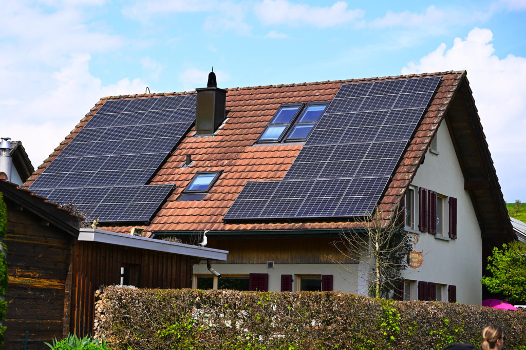 Einfamilienhaus mit Satteldach und roter Ziegeleindeckung, auf dem großflächig Photovoltaik-Module montiert sind, im Vordergrund eine Hecke