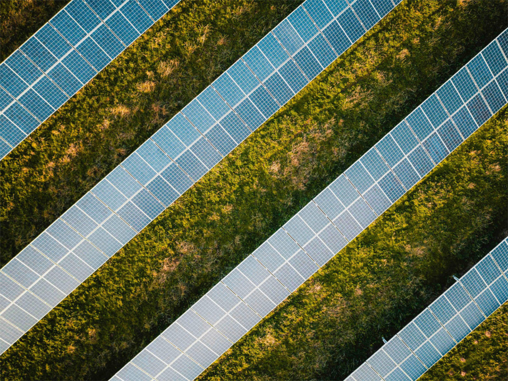 Luftaufnahme einer Freiflächen-Photovoltaikanlage: diagonal verlaufende Reihen von Solarmodulen auf einer grünen Wiese im Sonnenlicht