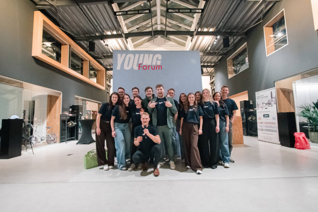 Gruppenfoto des YOUNG-forum-Teams in dunklen T-Shirts vor einer Wand mit YOUNG-forum-Logo in einer Industriehalle, vorne kniet ein Mann mit Mikrofon.