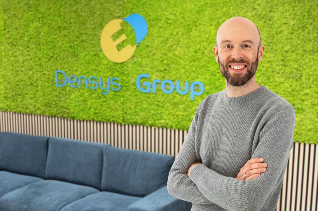 Steffen Binzel von Densys pv5 steht lächelnd mit verschränkten Armen vor einer Mooswand mit dem Logo der Densys Group, im Hintergrund ein blaues Sofa.