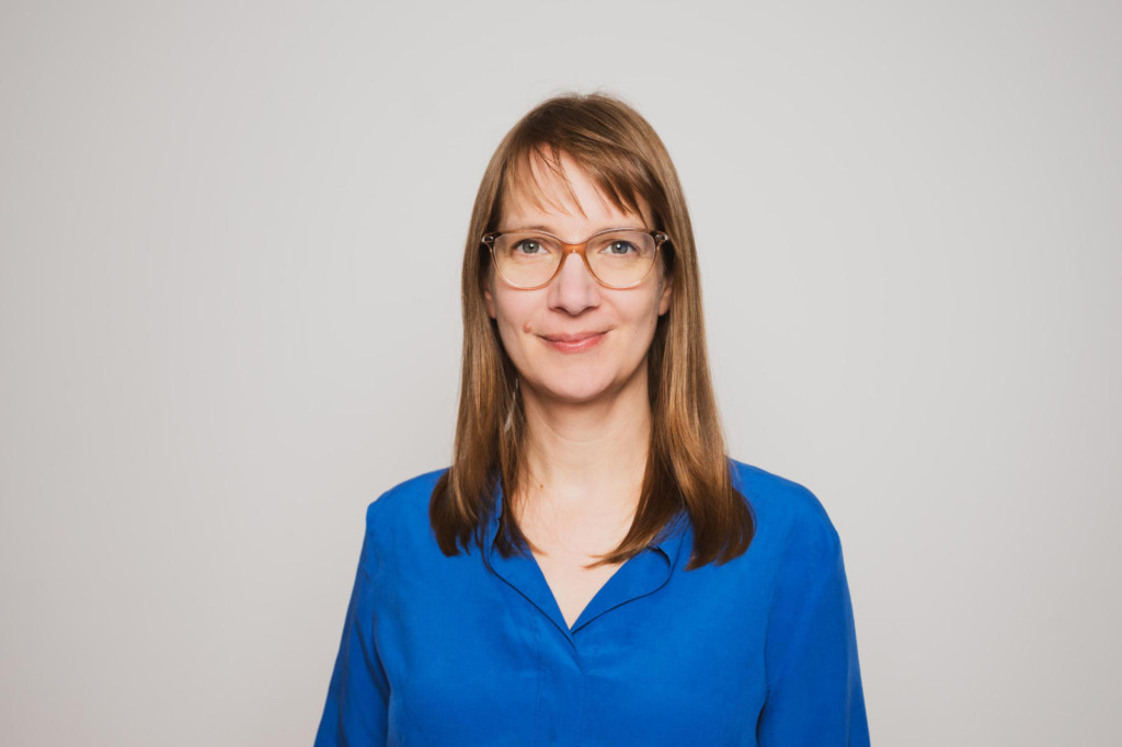Porträt von Kirsten Kelleher, neue Redaktionsleitung der SONNENENERGIE, mit Brille, schulterlangen braunen Haaren und blauer Bluse vor hellgrauem Hintergrund.