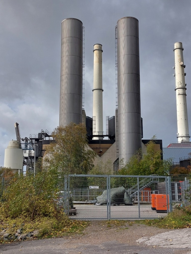 Kohlekraftwerk Wedel mit zwei hohen silbernen Schornsteinen und zwei weißen Schloten vor grauem Wolkenhimmel, im Vordergrund ein geschlossenes Gittertor.