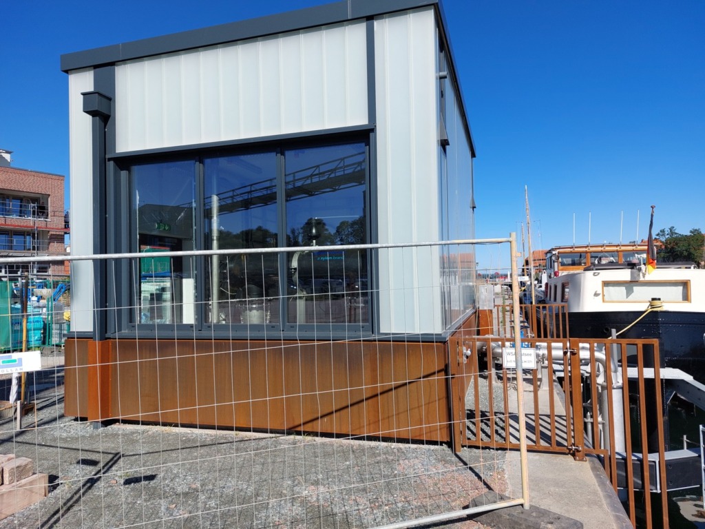 Pumpenhaus der Meerwasser-Wärmepumpe an der Kaikante in Neustadt in Holstein: weißer Flachbau mit Fensterfront, rechts Edelstahlrohre hinter einem Gittertor
